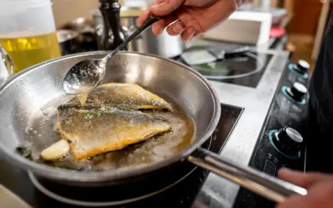 Un chef cocina pescado en una sartén de acero inoxidable, bañándolo con aceite caliente.