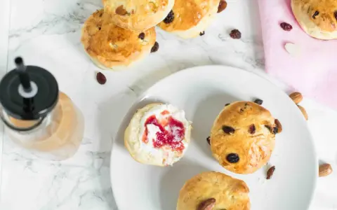Goldbraune Scones mit Rosinen und Mandeln, ein Scones Teig mit Marmelade und Sahne, daneben ein Getränk.
