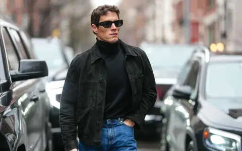Hombre con gafas de sol y chaqueta de gamuza negra sobre jersey de cuello alto, demostrando como tener estilo.