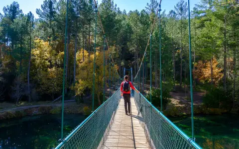 Senderista cruza puente colgante sobre el río, camino al Salto de Poveda y la Laguna de Taravilla. Bosque otoñal.
