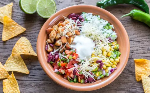 Bunter Taco Salat mit Hähnchen, Reis, Bohnen, Mais, Tomaten, Käse und Sauerrahm. Tortilla-Chips und Limetten daneben.