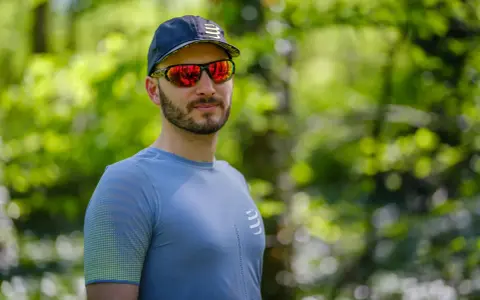 Mężczyzna w czapce i sportowych okularach przeciwsłonecznych, z odbiciem lasu w soczewkach.