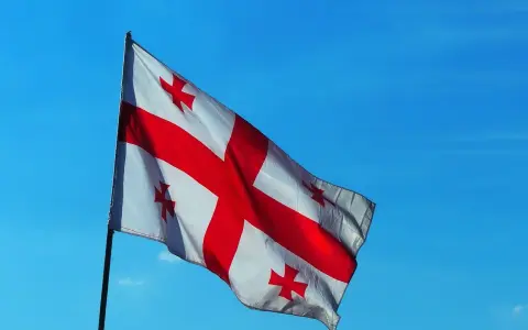 Flaga Gruzji powiewa na tle błękitnego nieba. Białe tło przecina czerwony krzyż, a w każdym z czterech pól znajduje się mniejszy czerwony krzyżyk.