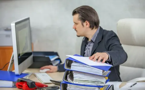 Mężczyzna w garniturze pracuje przy biurku, przeglądając dokumenty. Przed nim stos segregatorów, komputer i czerwony telefon. To obraz codziennego postępowania administracyjnego.