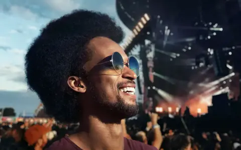 Un homme souriant avec des lunettes de soleil et des bouchons d'oreilles, profitant d'un concert en plein air. Le port des bouchons d'oreilles est obligatoire pour protéger son audition.