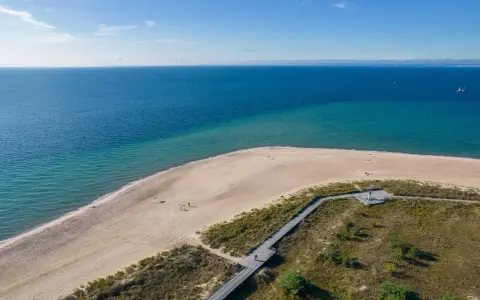 Piękne wydmy helskie nad turkusowym morzem. Drewniana kładka prowadzi przez zarośla na piaszczystą plażę, gdzie spacerują nieliczni turyści.
