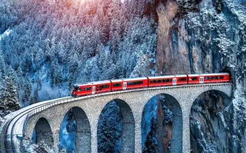 Un treno rosso attraversa un viadotto innevato, parte del suggestivo treno Bernina percorso, tra alberi coperti di neve e rocce imponenti.