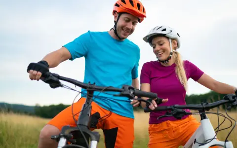 Uśmiechnięta para w kaskach i sportowych strojach, gotowa na przejażdżkę rowerami elektrycznymi. Wygodne akcesoria do roweru elektrycznego zapewniają komfort.