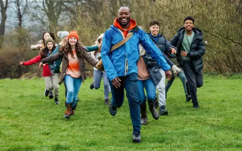 Lehrer und Schüler rennen lachend über eine grüne Wiese. Perfekt, um eine Klassenfahrt zu organisieren und Abenteuer zu erleben!