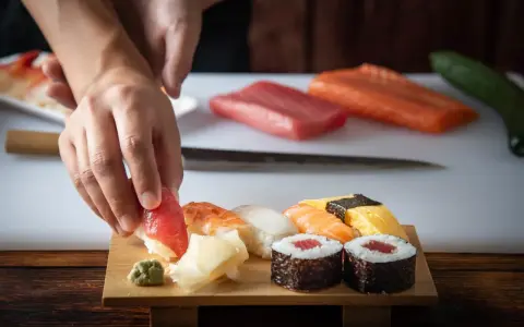 Ręce układają kawałek tuńczyka na ryżu, tworząc sushi. Na desce widać też krewetkę, kalmara, łososia i tamago. To inspiracja, jak zrobić sushi.