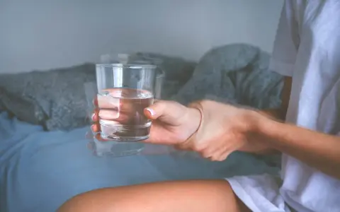 Dłoń z drżącym szkłem wody, sugerującym zwidy po alkoholu. Rozmyte kontury i niepewny chwyt.
