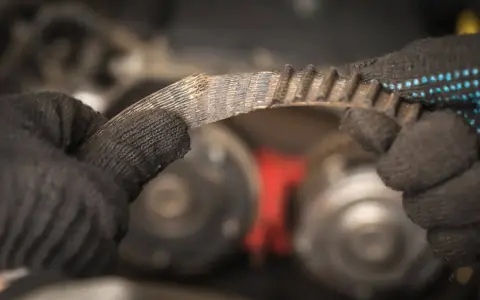 Zużyty, piszczący pasek napędowy w rękach mechanika. Widać postrzępione krawędzie i zużyte zęby, co sugeruje potrzebę wymiany.
