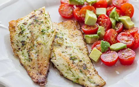 Dos filetes de lubina a la plancha con hierbas, acompañados de una ensalada fresca de tomates cherry y aguacate en cubos.