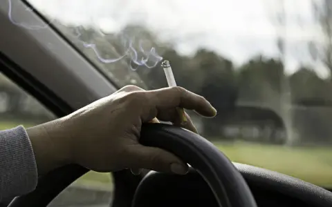 Mano sujetando un cigarrillo encendido sobre el volante. El humo se eleva mientras se puede fumar conduciendo.