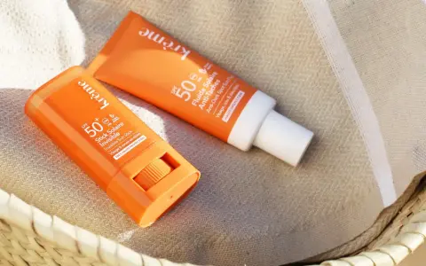 Deux produits solaires, un stick et un fluide, sont posés sur une serviette rayée. Le bouton du stick est visible, prêt à protéger du soleil.
