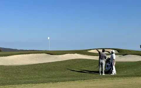 Zwei Golfer planen ihren nächsten Schlag auf dem Valderrama Golfplatz, mit einem Bunker und dem Fahnenmast im Hintergrund.