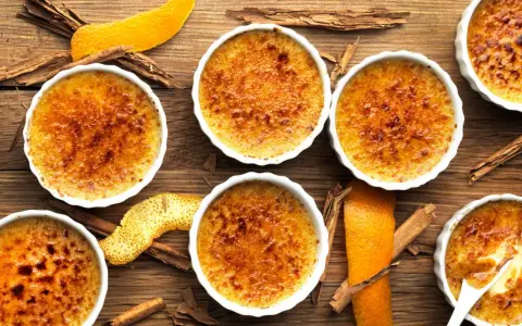 Flera krämiga crema catalana i vita skålar, toppade med karamelliserat socker. Apelsinskal och kanelstänger ger en härlig doft.