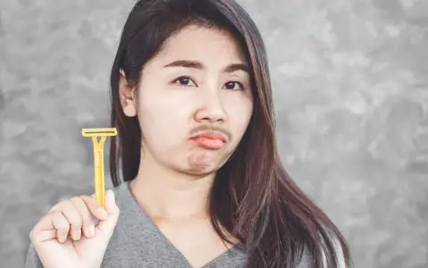 Mujer con expresión de disgusto sostiene una cuchilla amarilla, con pelos en la barbilla simulando bigote.