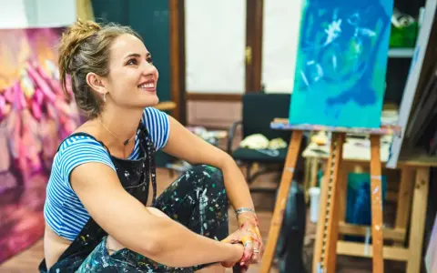 Une artiste rêveuse, les mains tachées de peinture, s'interroge sur comment devenir artisan d'art. Son atelier est rempli de toiles colorées.