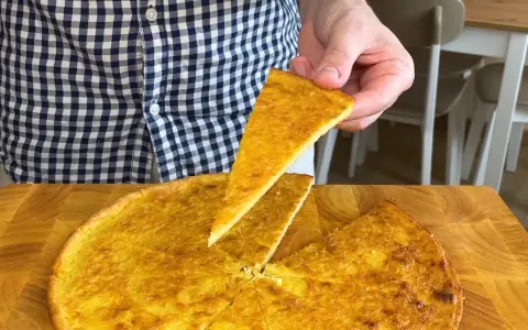 Una fetta di farinata di ceci dorata e croccante viene sollevata da una teglia rotonda su un tagliere di legno.