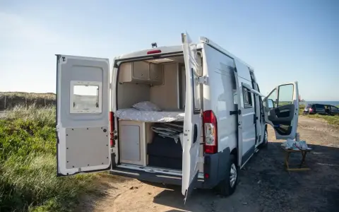 Biały kamper z otwartymi drzwiami, ukazujący łóżko i schowki. Idealne miejsce na życie w kamperze nad morzem.