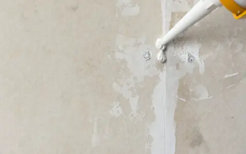 Nakładanie białego akrylu na pęknięcia i nierówności ściany, przygotowanie do malowania.