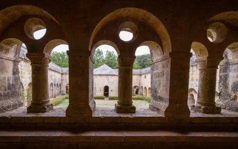 Vue à travers les arches de l'abbaye du Thoronet, un cloître en pierre avec un jardin verdoyant et des bâtiments anciens.