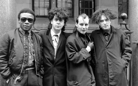 Vier Männer in dunkler Kleidung, Post-Punk-Ästhetik, stehen vor einem Gebäude.