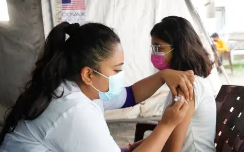 En sjuksköterska ger en vaccination i Nepal. Bakom syns en banderoll med texten COVAX och en amerikansk flagga.