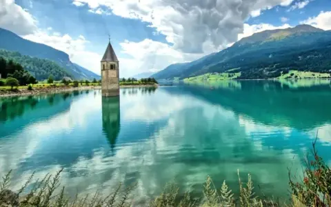 Il lago del campanile, con la sua torre sommersa, riflette montagne verdi e un cielo nuvoloso.