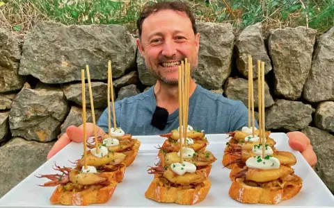 Hombre sonriente presenta cazuelitas de aperitivo con calamares y alioli sobre pan tostado, decoradas con brochetas.