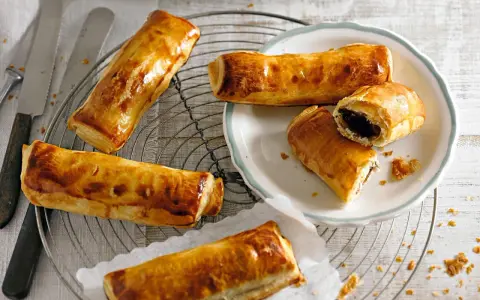Goldbraune, knusprige Pain au Chocolat auf einem Gitter, einige auf einem Teller, einer aufgeschnitten.