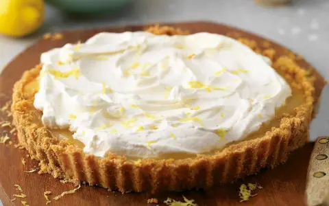 A lemon tart topped with whipped cream and lemon zest on a wooden board.
