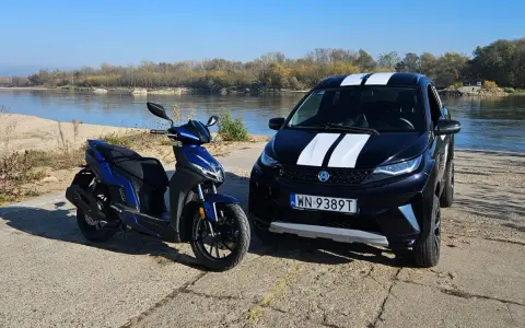 Niebieski skuter i czarne aixam auto z białymi pasami stoją nad rzeką.