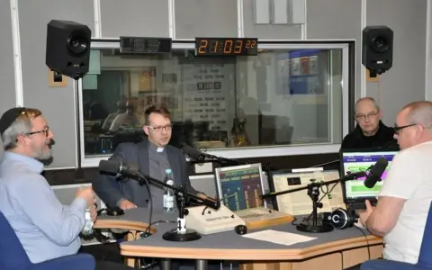 W studiu radiowym czterech mężczyzn, w tym ksiądz i osoba w jarmułce, rozmawia przy mikrofonach. W tle widać zegar i logo RDC.