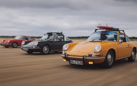 Trzy klasyczne Porsche jadące po plaży. Zdjecie w ruchu, z deskami surfingowymi na dachu jednego z nich.