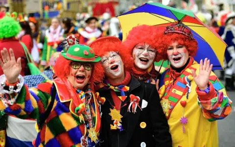 Vier Clowns mit roten Perücken und bunter Kleidung lachen und winken. Ein bunter Regenschirm ist über ihnen. Hier wird kein Karneval gefeiert, sondern ausgelassen gefeiert!