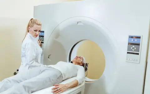 Kobieta w białej bluzie leży na łóżku rezonansu magnetycznego. Technik w białym fartuchu obsługuje panel sterowania.