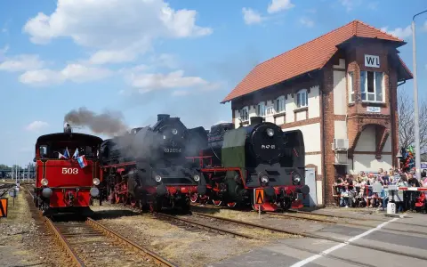 Parowóz 503 i dwa czarne parowozy na torach przed budynkiem z napisem WL. Muzeum Kolejnictwa Wolsztyn.