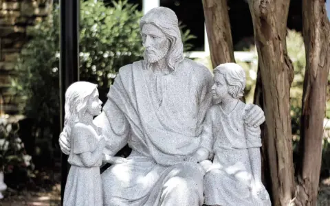 Steinfigur von Jesus, der zwei Kinder umarmt. Er wirkt wie ein liebevoller Gottvater, der seine Schützlinge beschützt.