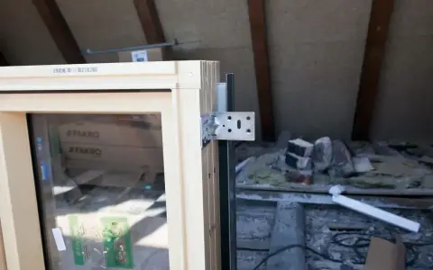 Drewniane okno z metalowym kątownikiem, gotowe do obróbki po montażu. W tle widać materiały budowlane i drewniane belki poddasza.
