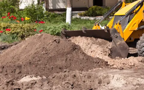 Koparka z łyżką zasypuje ziemię, przygotowując teren pod budowę. W tle kwitnące tulipany.