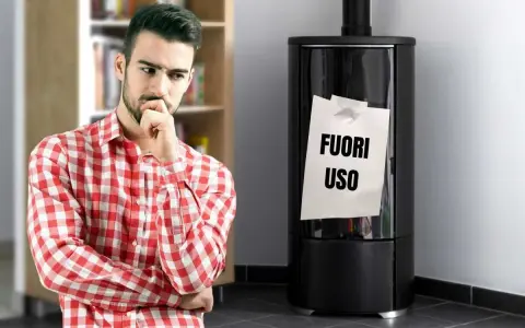 Uomo pensieroso guarda stufa spenta con foglio "FUORI USO". Necessaria riparazione stufe per il freddo.