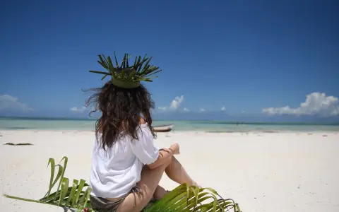 Kobieta w koronie z liści siedzi na plaży, marząc o tym, jak dolecieć na Zanzibar. Błękitne niebo i turkusowa woda zapraszają do podróży.