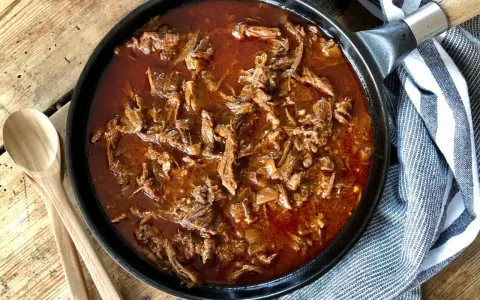 En gjutjärnspanna fylld med saftig, långkokt pulled beef i en mustig sås. Två träskedar ligger bredvid, redo att servera.