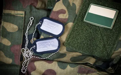 Dwa nieśmiertelniki na mundurze moro. Jeden z nich zawiera napis "MILITARIA.PL SHOOTING & OUTDOOR".