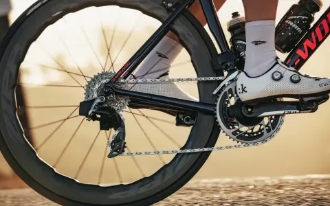 Close-up of a cyclist's shoe and drivetrain, showing the chain, gears, and crankset. The focus is on the power transfer, hinting at the cycling watts being generated.