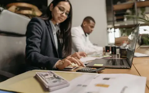 Kobieta w okularach liczy na kalkulatorze, obok stos banknotów i laptop.
