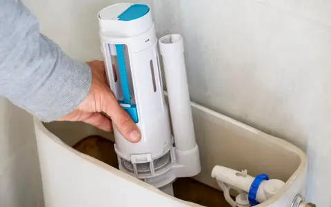 Mano instalando el mecanismo de descarga, una de las partes de una cisterna de baño. Se ve el flotador y la tubería de llenado.