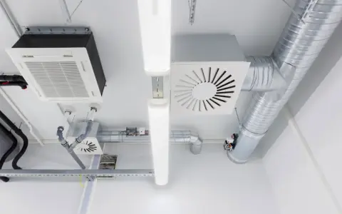 Detalles de sistemas de ventilación y extracción de aire, incluyendo conductos metálicos, unidades de aire acondicionado y luces fluorescentes en un techo.